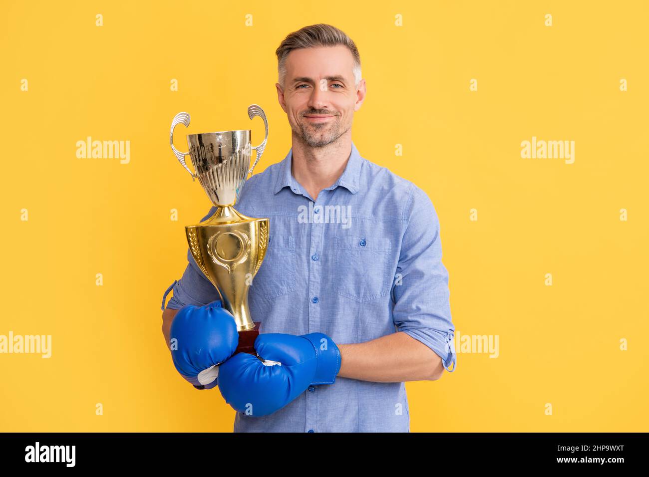 smiling adult man in boxing gloves hold golden cup on yellow background, victory Stock Photo