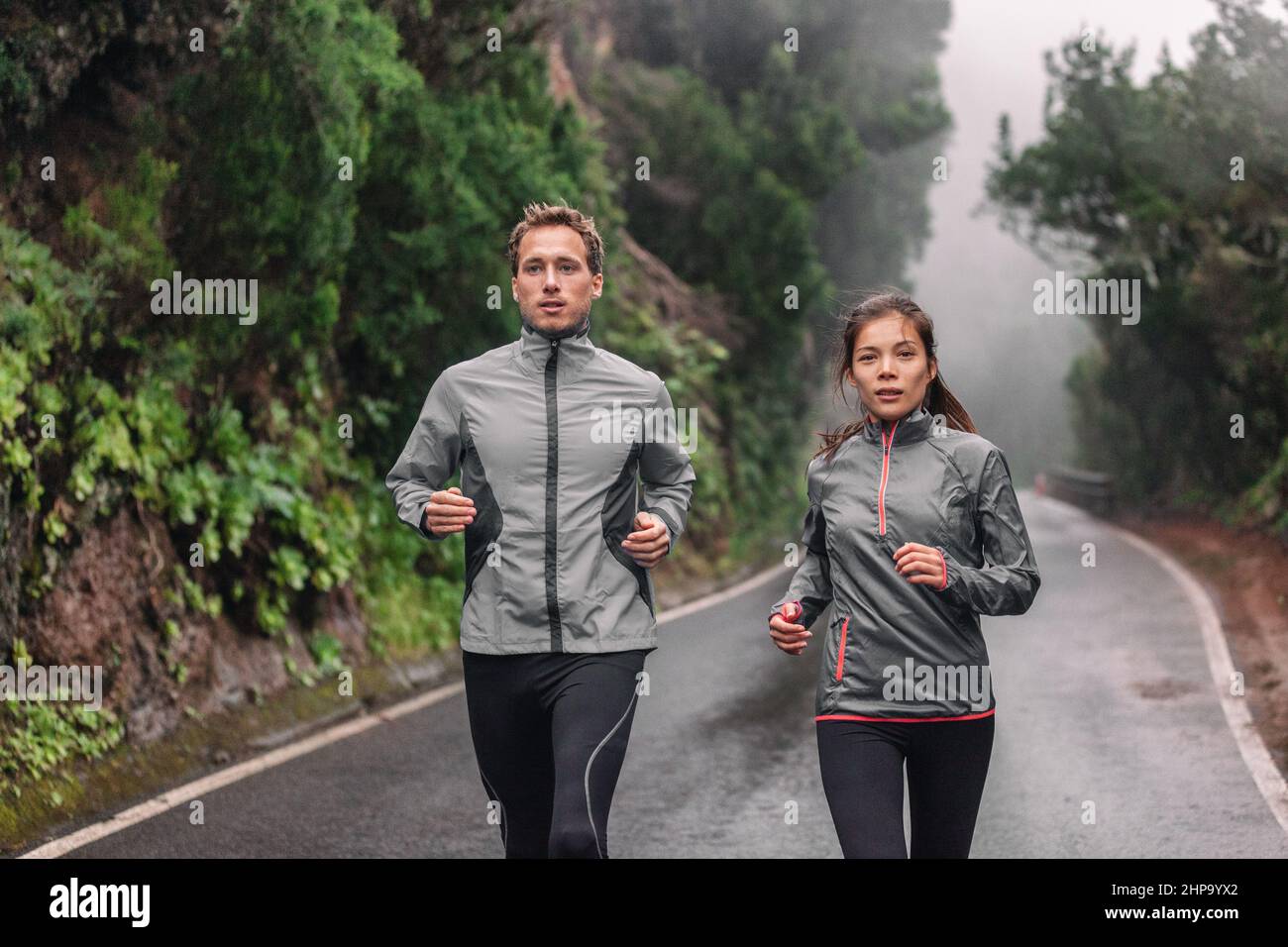 Running couple on wet park trail jogging in rain wearing sportswear jackets in cold weather. Asian woman, Caucasian man athletes training cardio Stock Photo