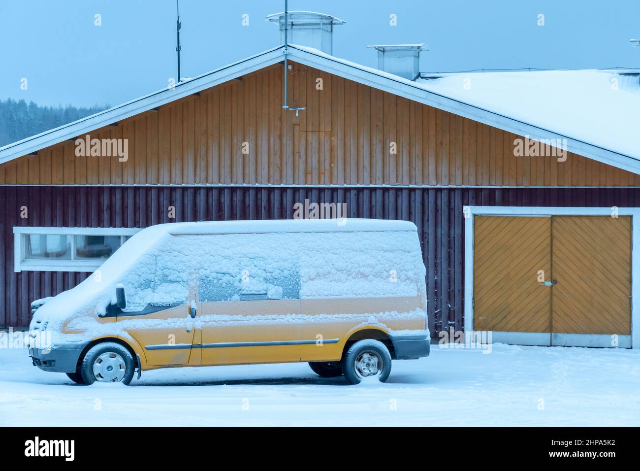 Yellow Ford Transit van covered in snow in front of a colored door of barn in Finland Stock Photo