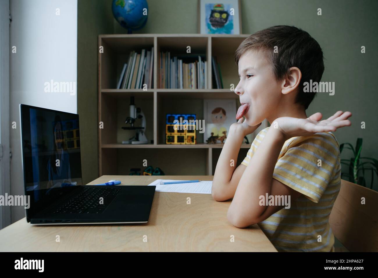 8 years old boy sit by desk with laptop and sticking out his tongue and