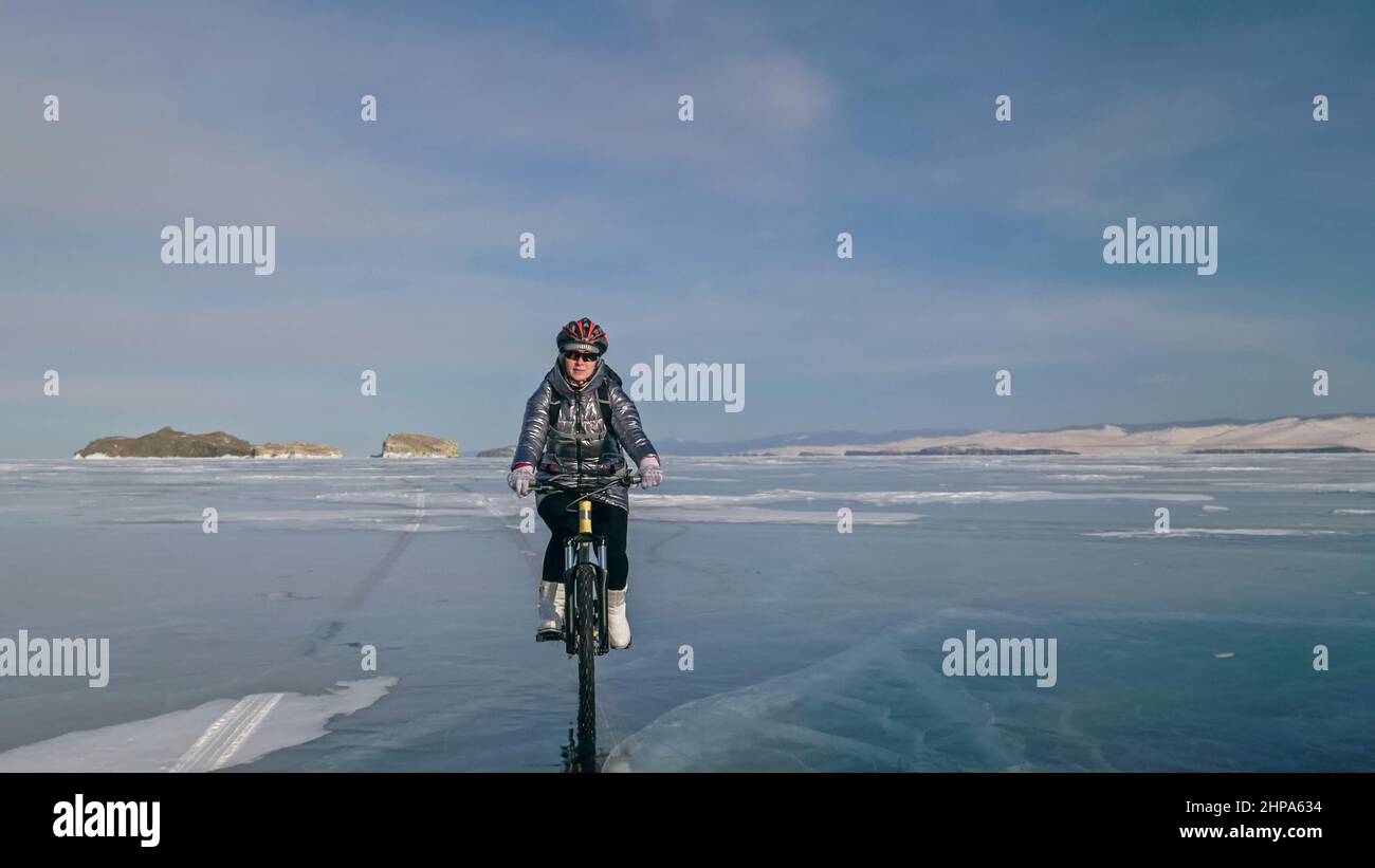 Woman is riding bicycle on the ice. Girl is dressed in a silvery down jacket, cycling backpack and helmet. Ice of the frozen Lake Baikal. Tires on bik Stock Photo