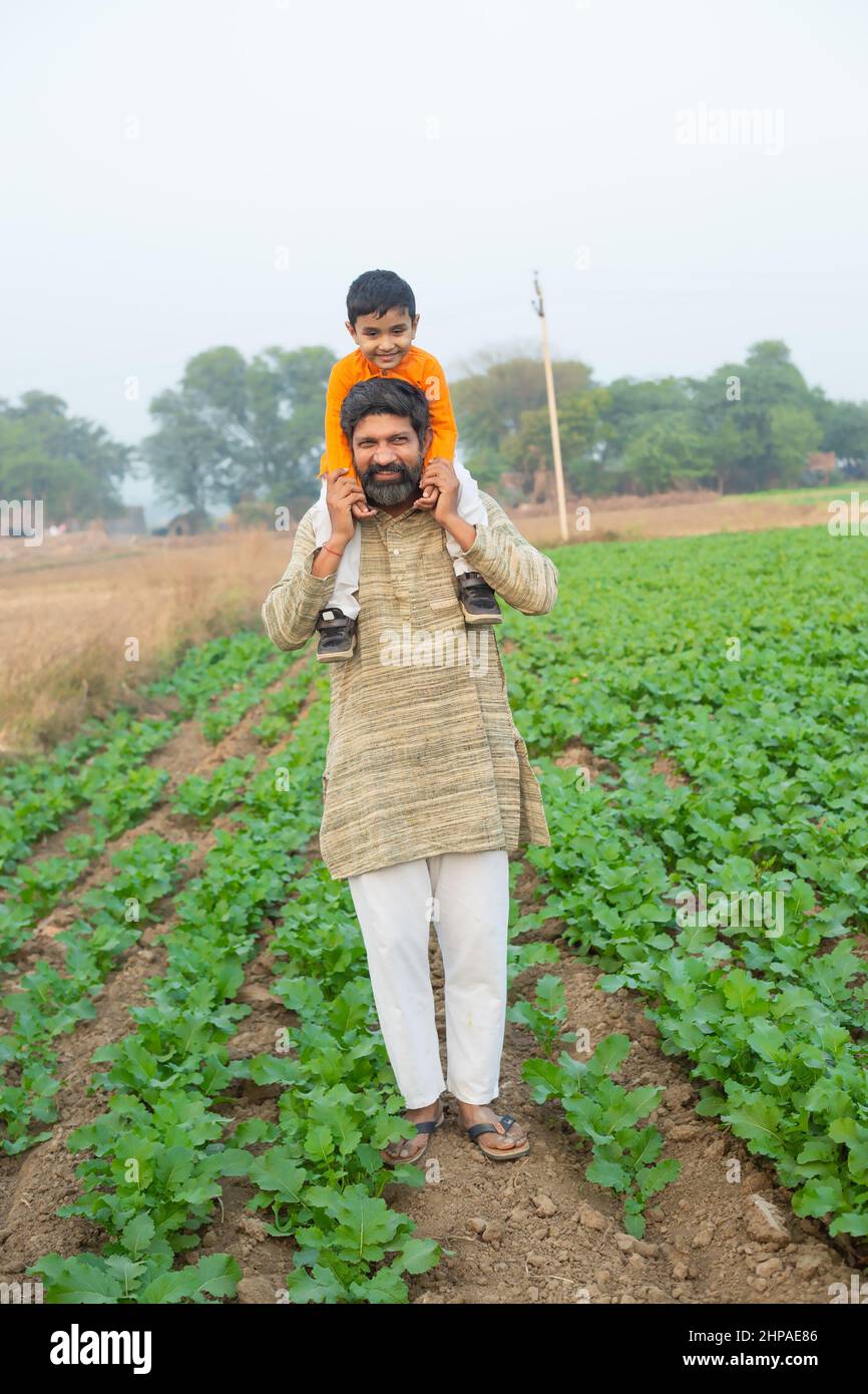 Happy Indian son sitting on father shoulder having fun in agriculture field, spend leisure time together in farm, boy child enjoy childhood with man o Stock Photo