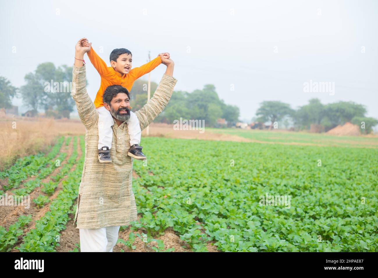 Happy Indian son sitting on father shoulder having fun in agriculture field, spend leisure time together in farm, boy child enjoy childhood with man o Stock Photo