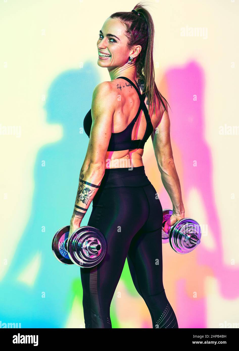 Sporty smiling woman working out with dumbbells. Photo of muscular woman in black sportswear on white background with effect of rgb colors shadows. Sp Stock Photo