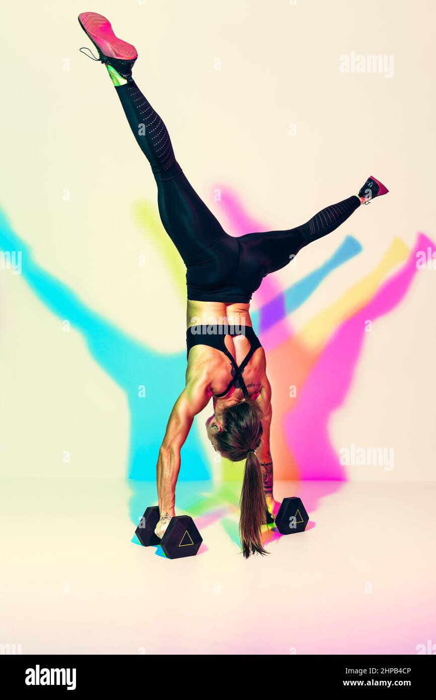 Strong woman does exercise handstand pushups on dumbbells. Photo of athletic woman in black sportswear on white background with effect of rgb colors s Stock Photo