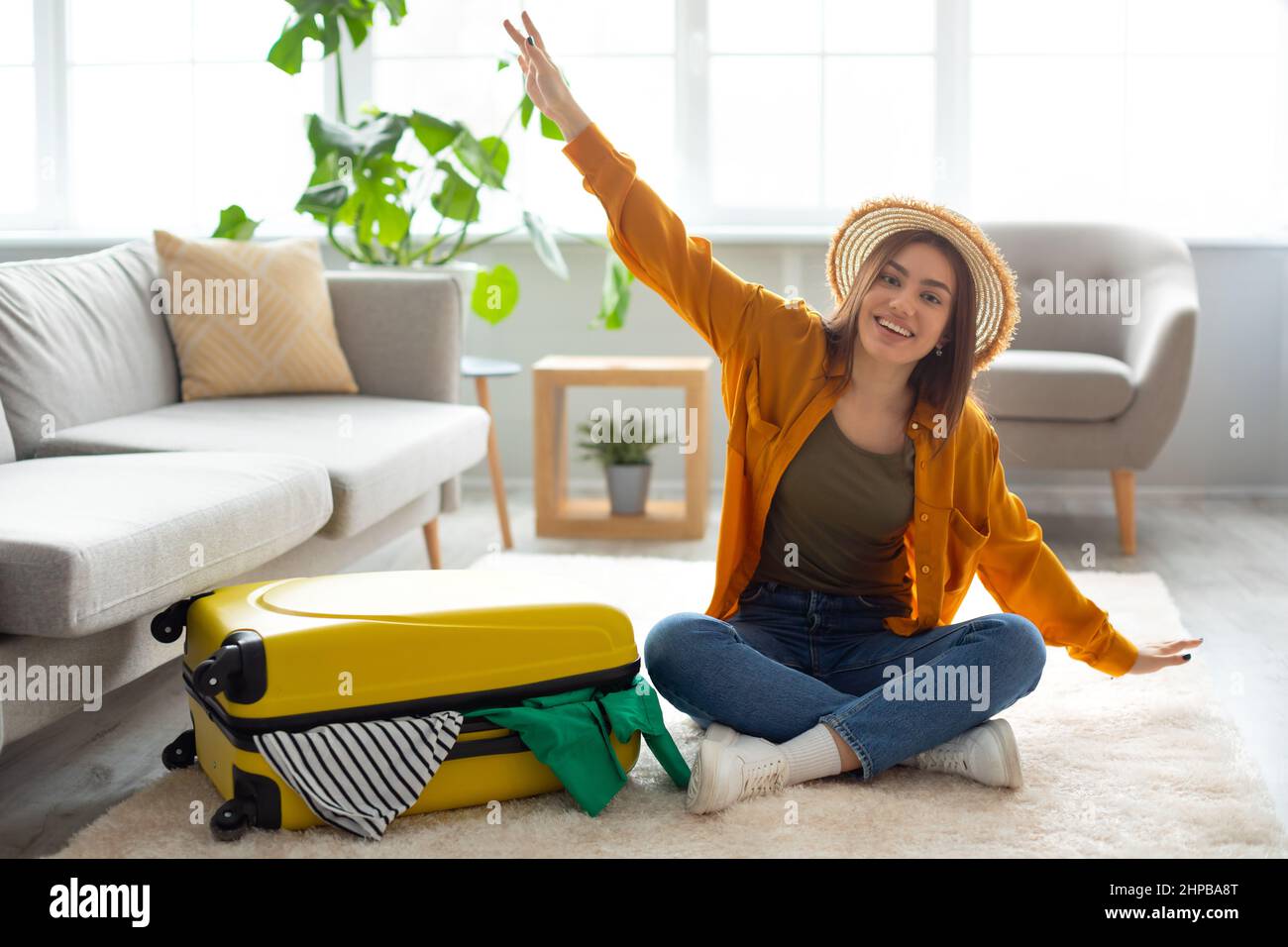 Cheery young lady sitting on floor near packed suitcase, making airplane gesture, feeling happy about abroad vacation at home, copy space. Traveling o Stock Photo