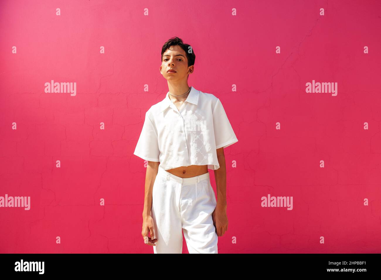 Self-confident queer boy standing against a pink background. Young teenage boy looking at the camera while standing in a studio. Confident gay teenage Stock Photo