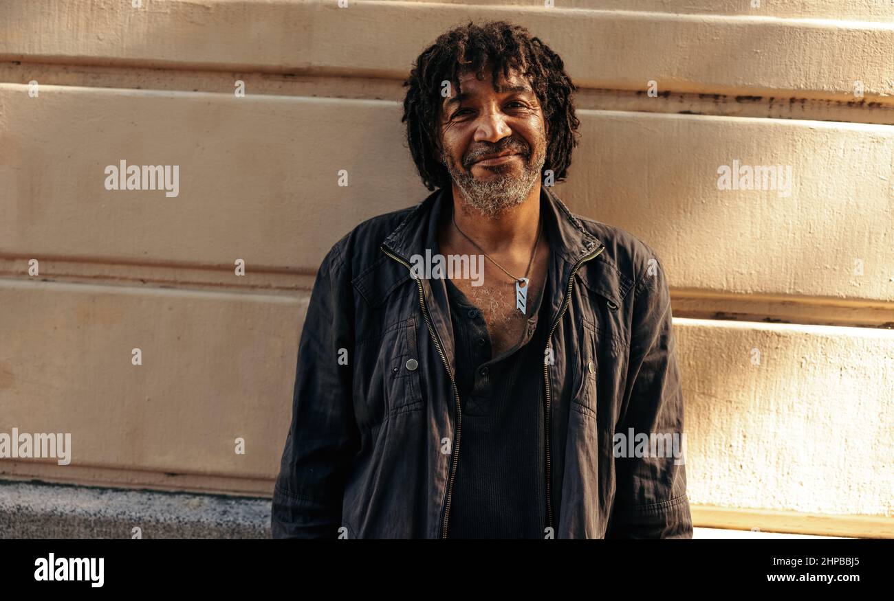 Cheerful homeless man smiling at the camera with confidence while standing alone during the day. Self-confident underprivileged man standing against a Stock Photo