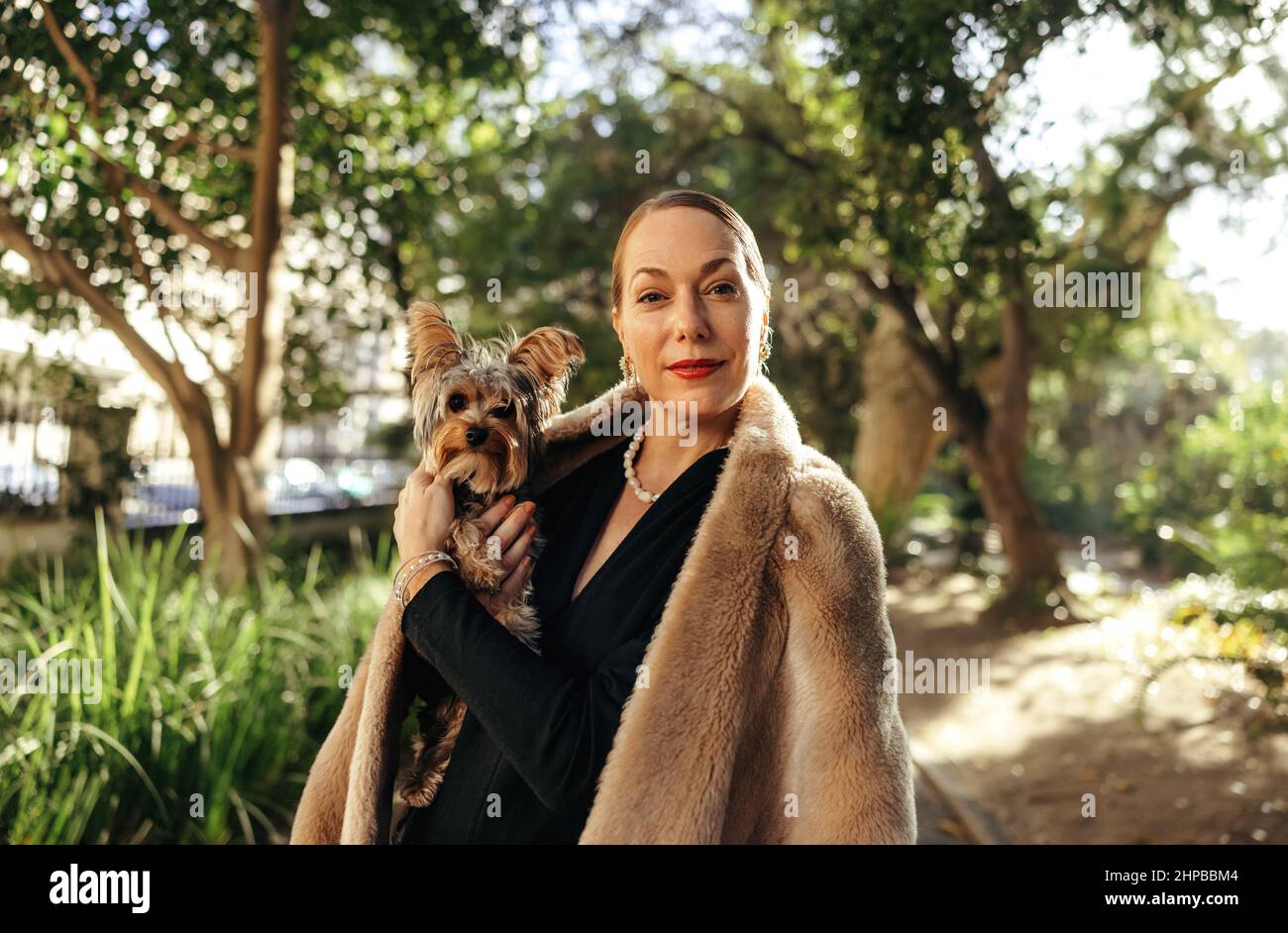High class woman looking at the camera while holding her puppy outdoors. Middle aged posh woman standing alone in a park during the day. Female pet ow Stock Photo