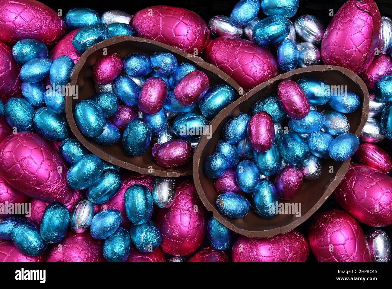Pile of different sizes of colourful foil wrapped chocolate easter eggs in pink, red, silver and blue with two halves of a large chocolate egg. Stock Photo