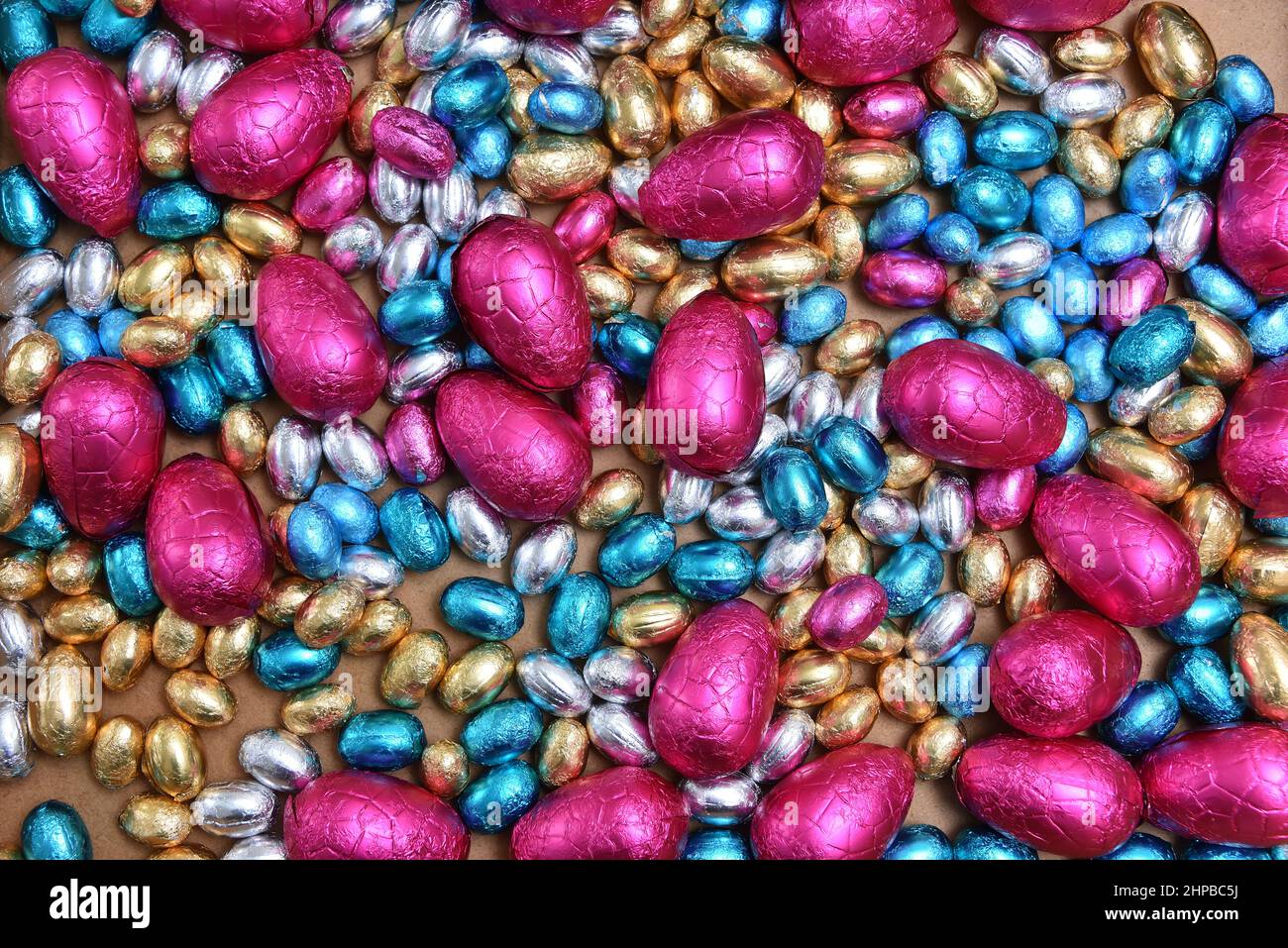 Small pink, blue, silver and gold foil wrapped chocolate easter eggs, with large pink eggs against a pale wood background. Stock Photo