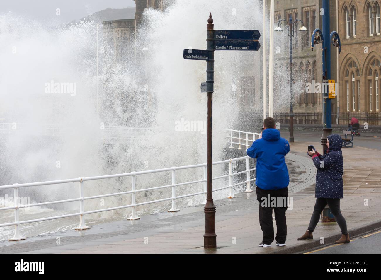 Aberystwyth, Ceredigion, Wales, UK. 20th February 2022  UK Weather: People brave the windy weather as they take photos while walking along the promenade, as Stormy weather continues along the west coast of Aberystwyth. with combining high tide, bringing rough seas crashing against the promenade and sea defences. © Ian Jones/Alamy Live News Stock Photo