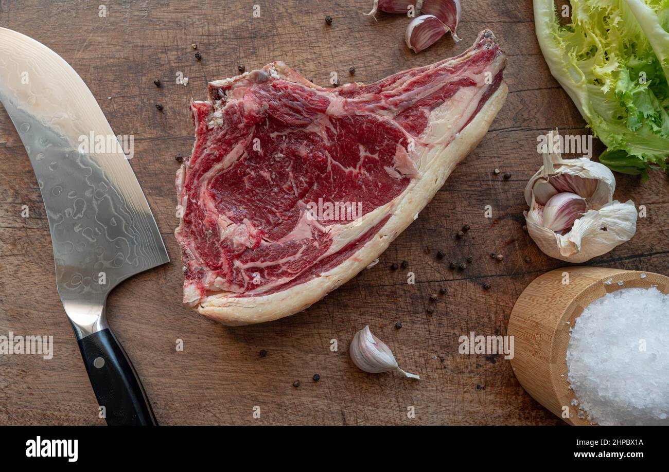 Delicious dry aged raw rubia gallega beef steak ready-to-be-cooked on a wooden board with knife, sea salt flakes, pepper, garlic, green salad Stock Photo