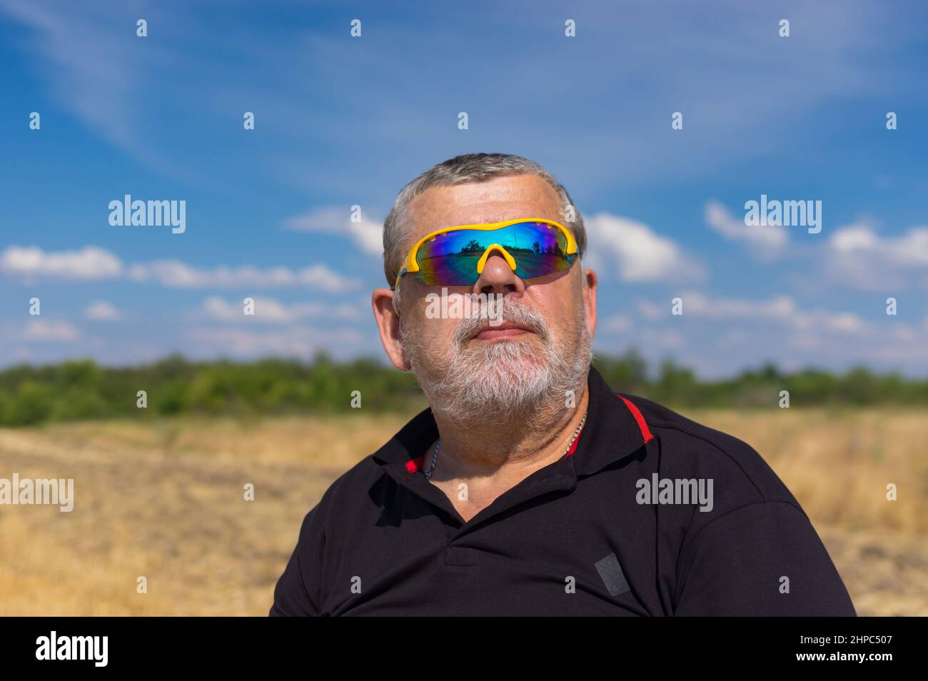 Outdoor portrait of a bearded senior in sunglasses against blue cloudy sky and agricultural field Stock Photo