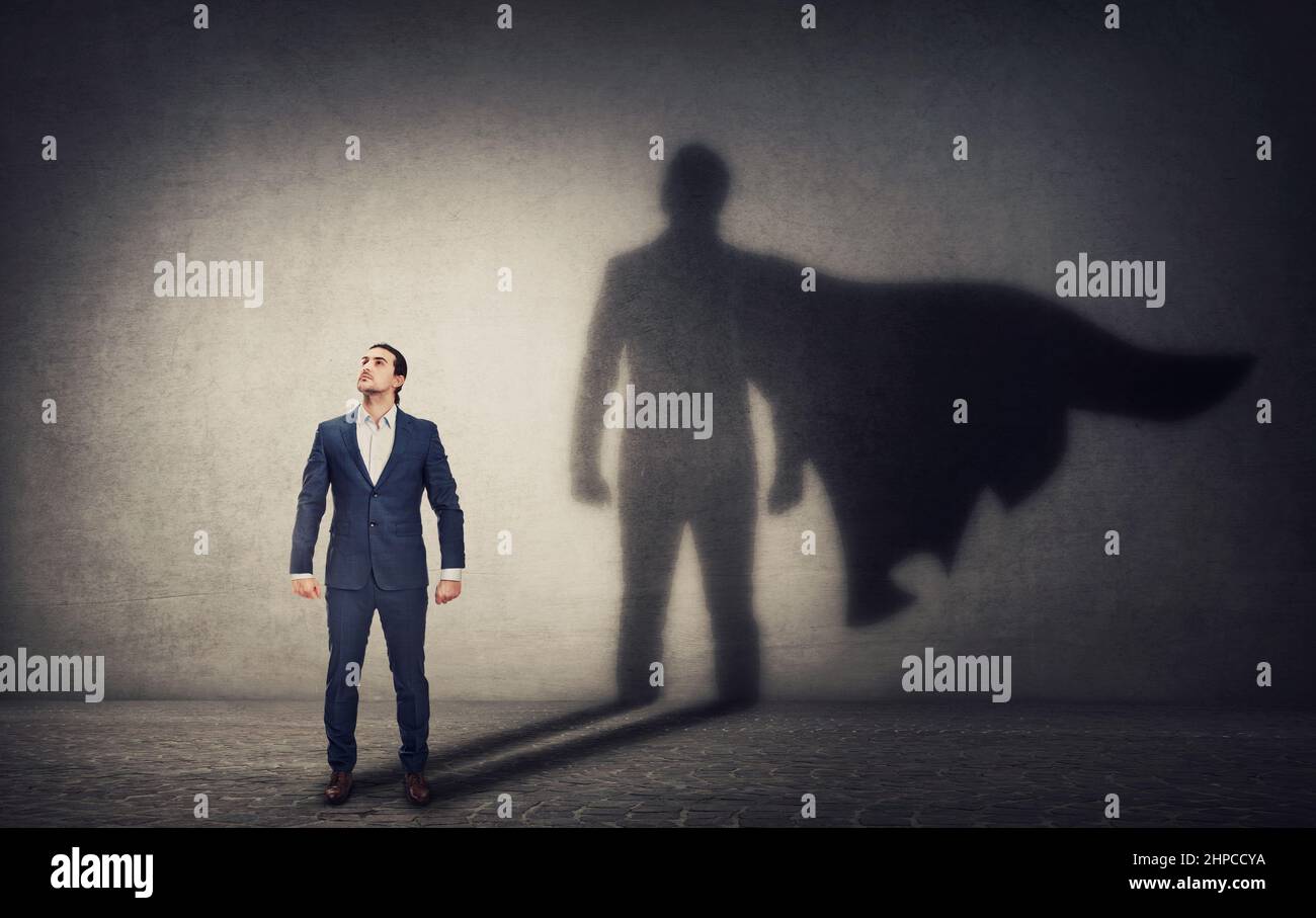 Determined businessman stands confident in a hero stance and casting a brave superhero shadow on the wall behind. Business leadership and motivation c Stock Photo