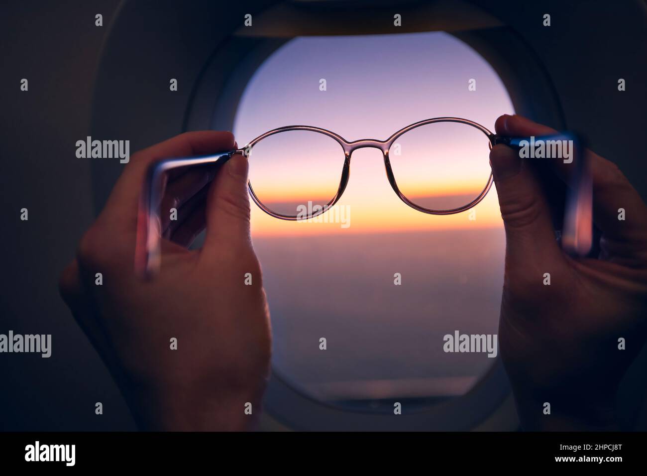 Hand of passenger holding eyeglasses against airplane window during colorful sunset. Themes travel, eyesight and vision. Stock Photo