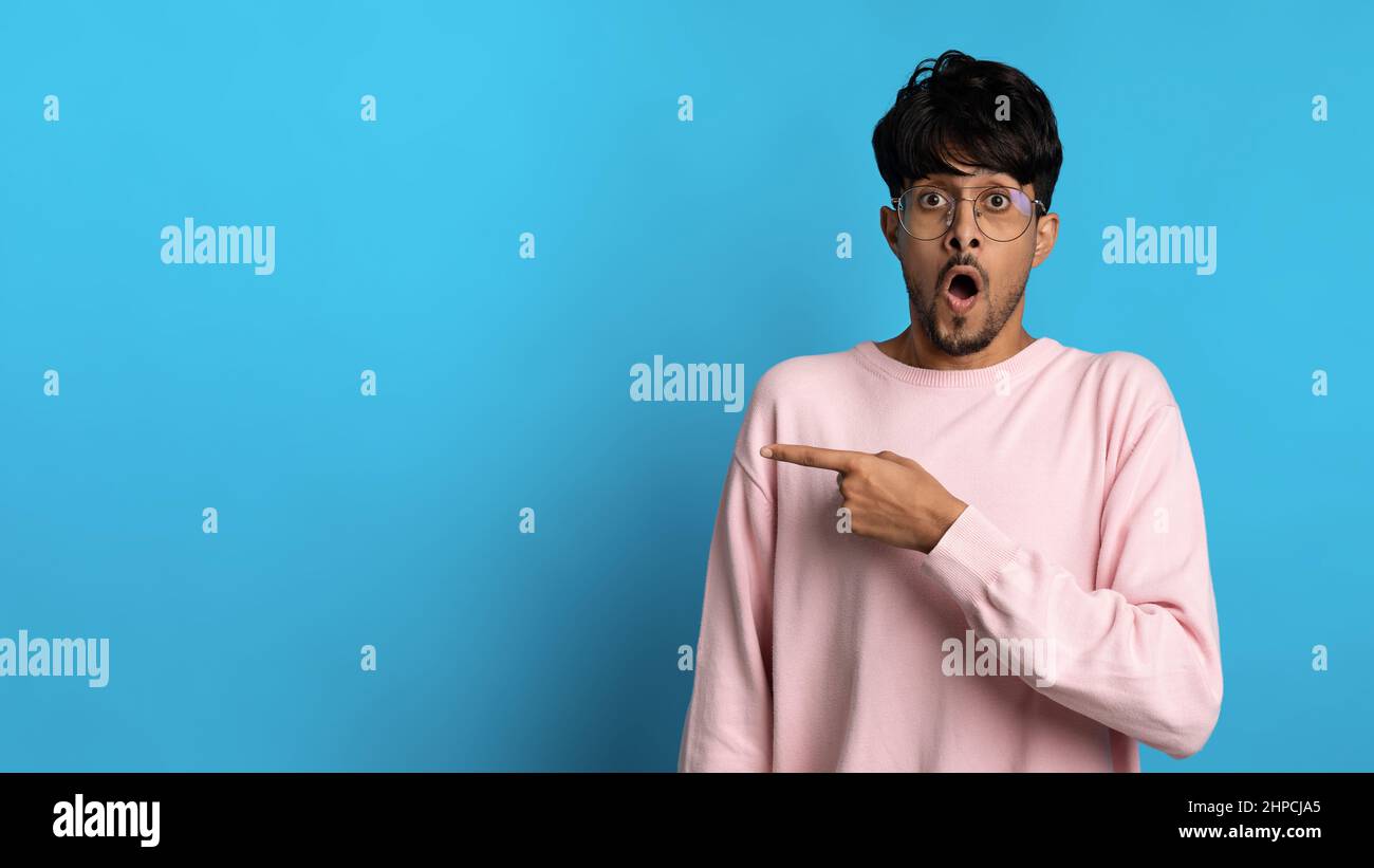 Shocked bearded indian guy wearing eyeglasses and pink sweatshirt pointing at copy space for text on blue studio background, showing shocking advertis Stock Photo