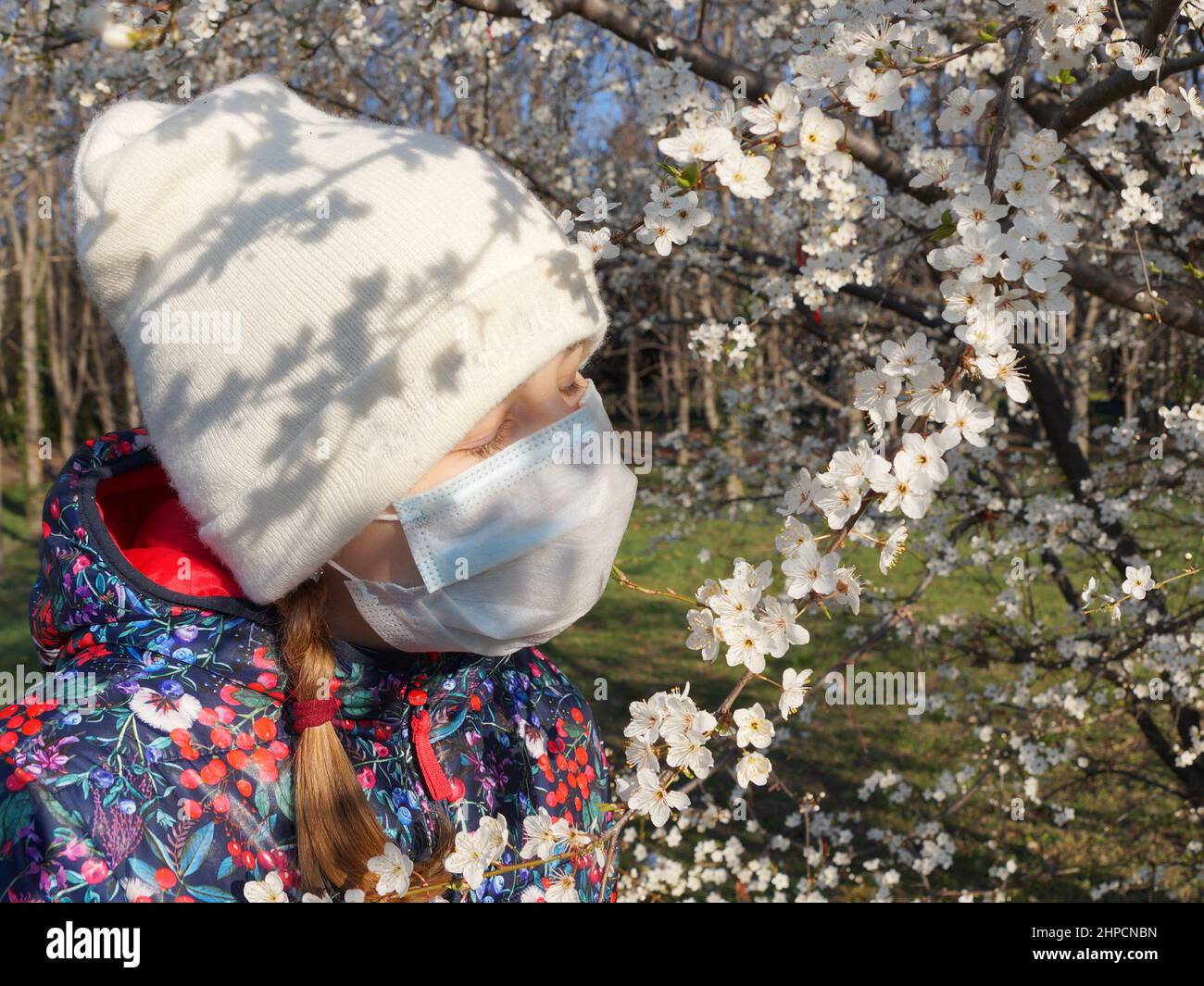 a girl in a hat and a medical mask sniffs a flowering branch in the park. Stock Photo