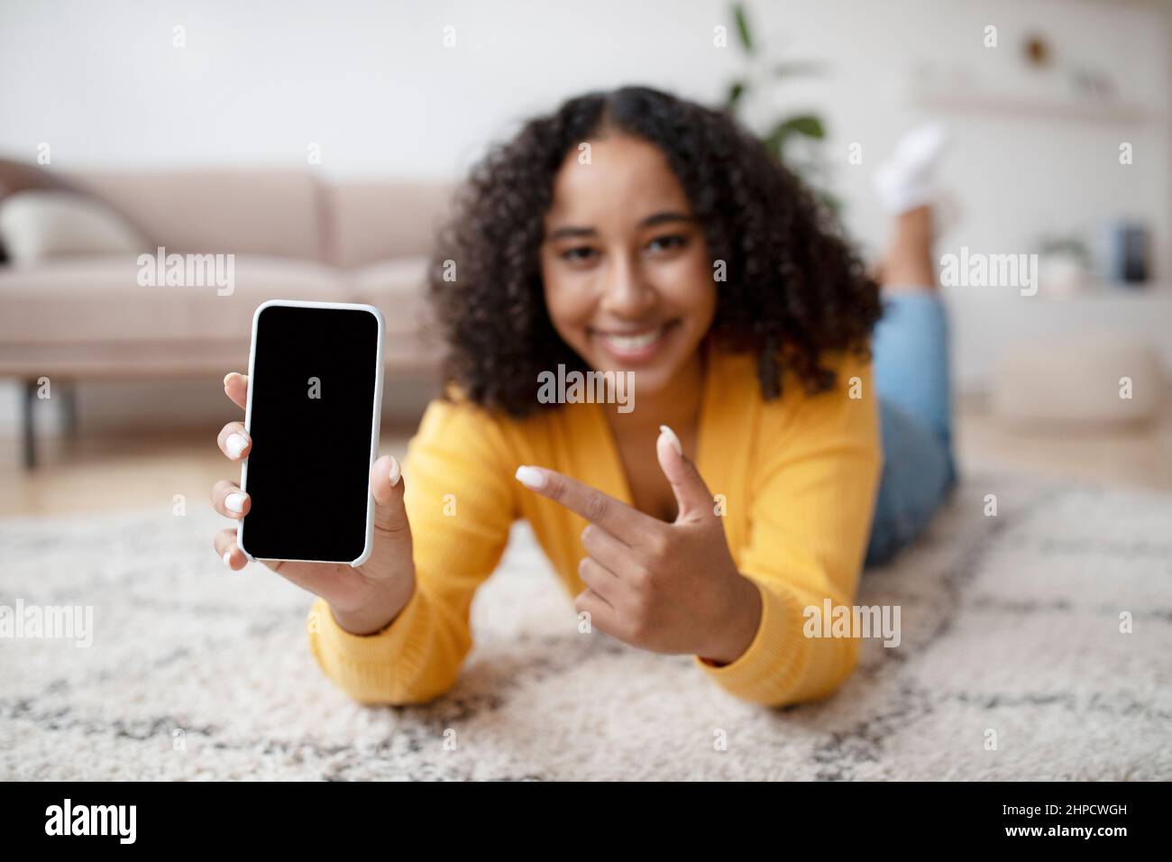 Happy young African American woman pointing at smartphone with empty screen, lying on floor at home, demonstrating mockup for website or mobile app. S Stock Photo