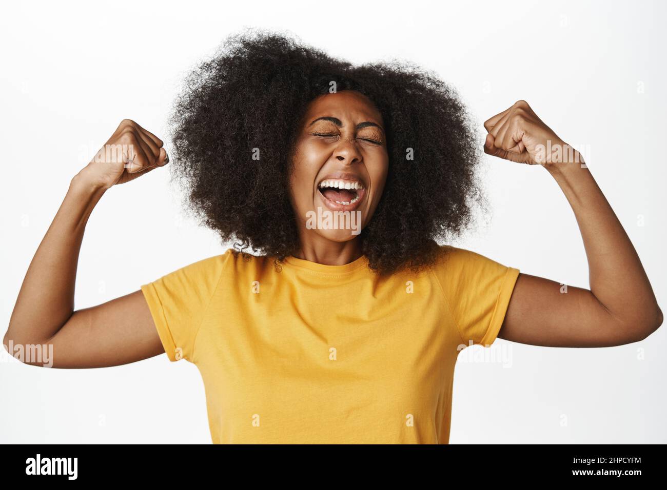 Close up portrait of enthusiastic black woman shouts with joy, flexing biceps and muscles, feeling strong and powerful, standing over white background Stock Photo