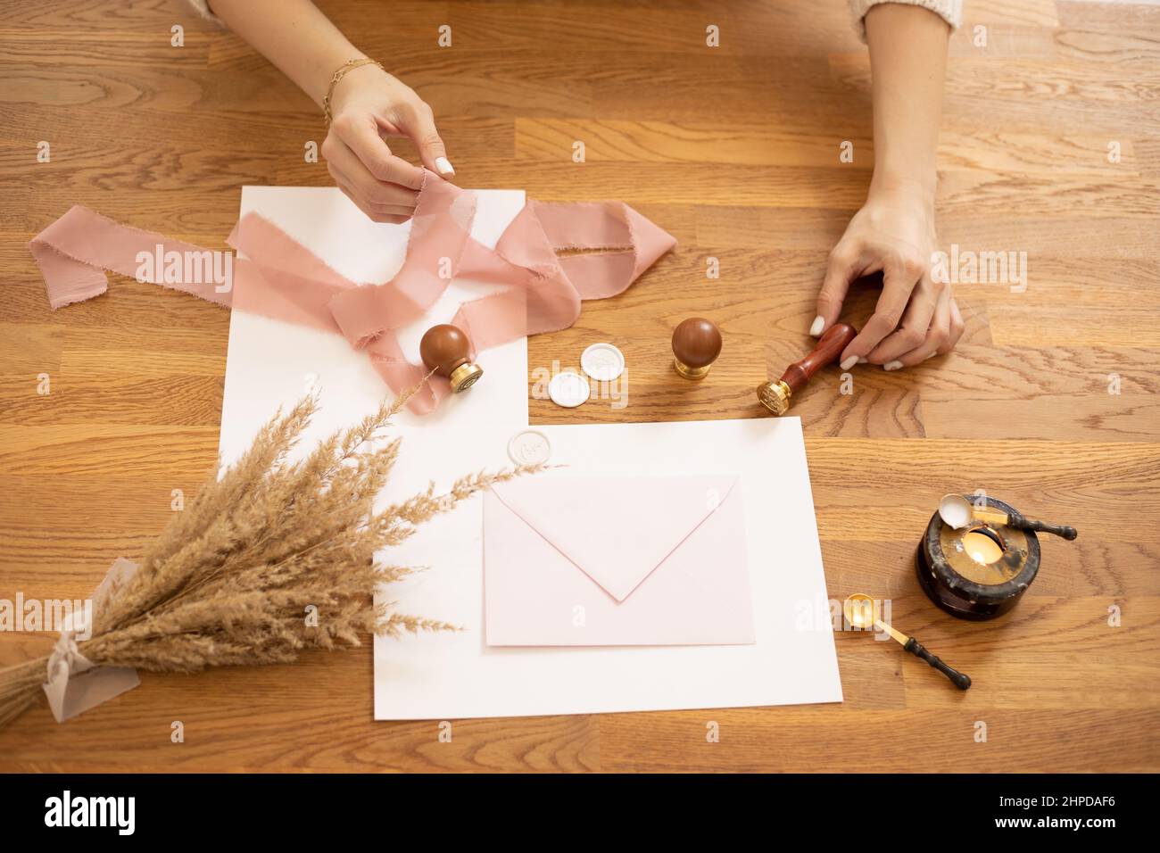 Cropped photo of woman hands making greeting envelope craft, decorate with pink ribbon and seal-wax. Cereals on desk.  Stock Photo