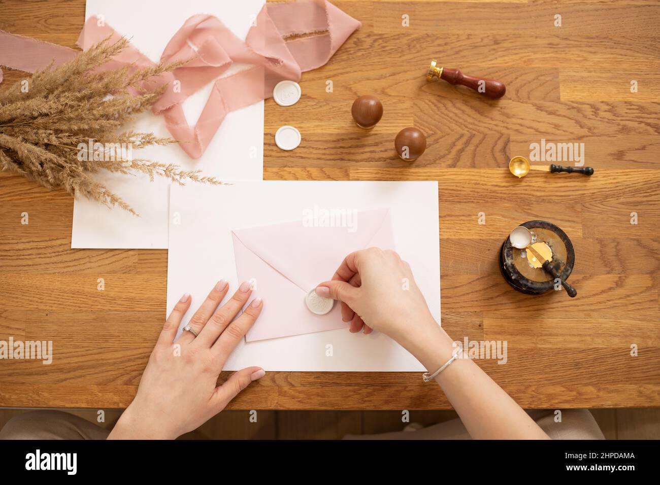 Cropped photo of woman hands making greeting envelope craft, decorate with pink ribbon and stamping. Flowers on desk.  Stock Photo