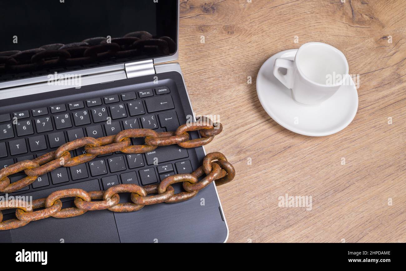 Computer keyboard locked by old rusty chain with reflection on display. Laptop detail and empty coffee cup on wooden desk. Bans and threats to freedom. Stock Photo