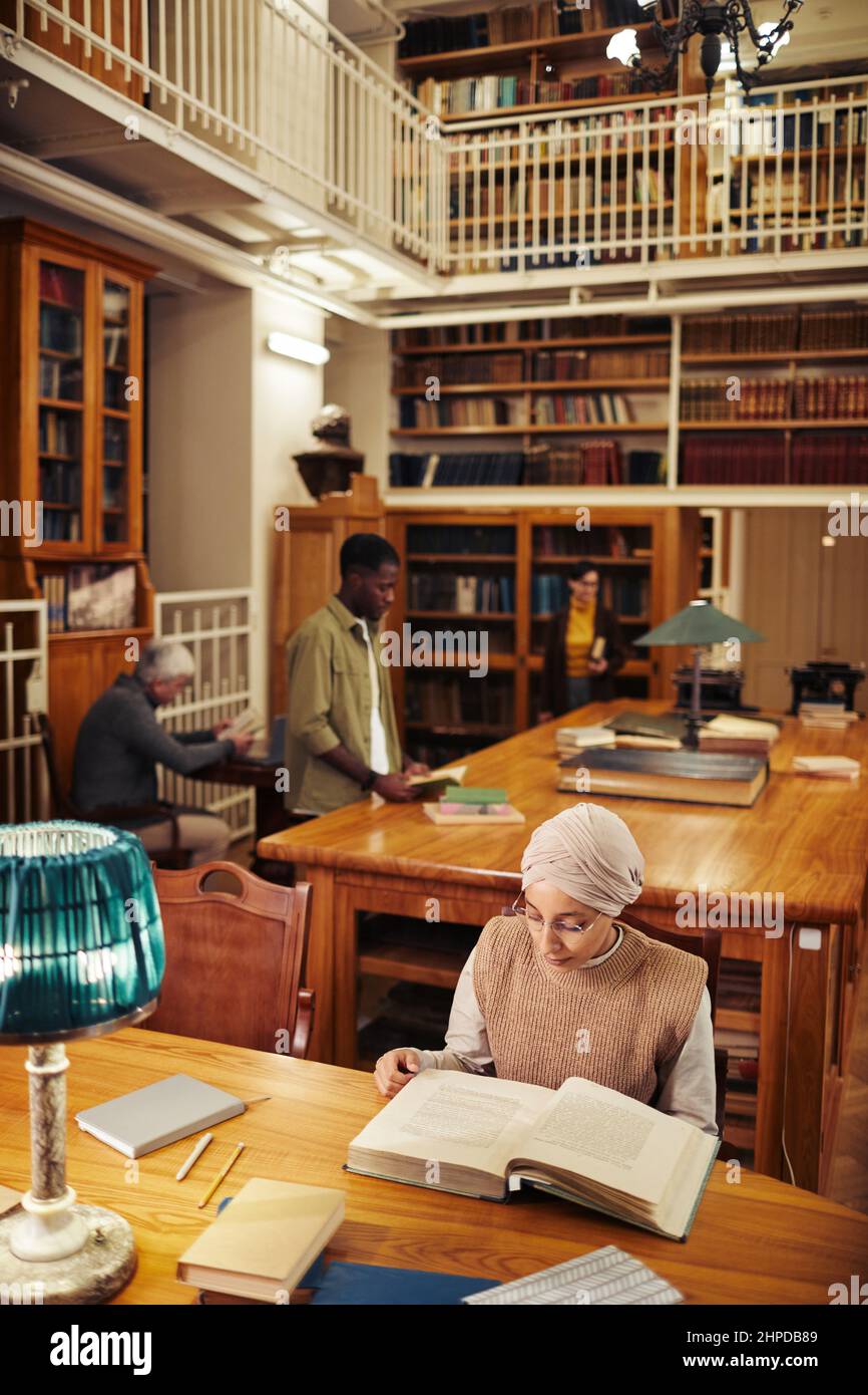 Vertical wide angle shot of classic college library with diverse group of people, focus on young woman wearing head covering and reading book Stock Photo
