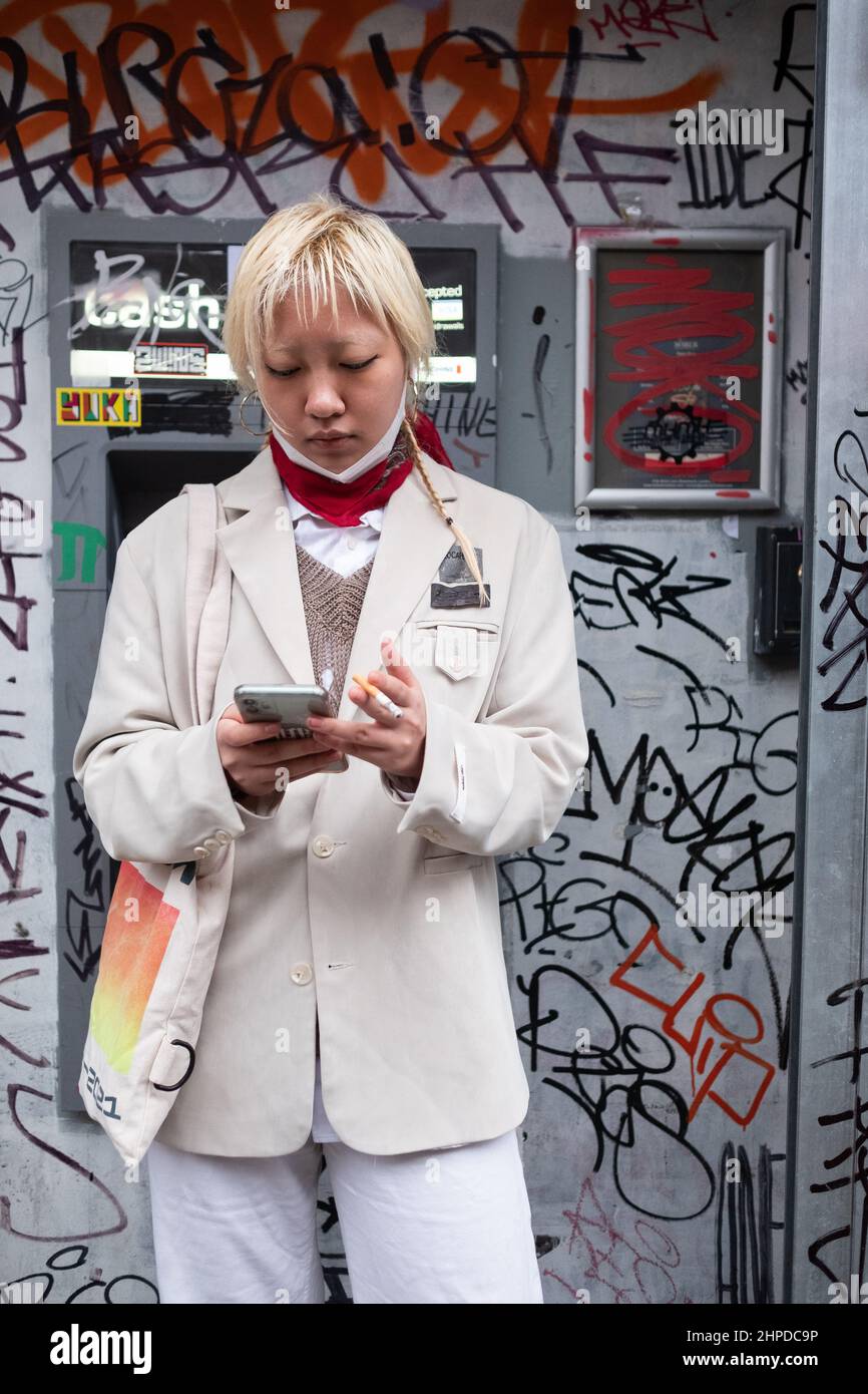An Asian female checks her phone whilst smoking a cigarette. Behind is a heavily graffitied cash point. Image taken in East Londons Brick Lane. Stock Photo