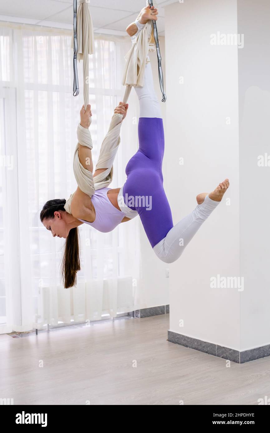 brunette woman 25 years old is hanging upside down using hammock for yoga. Lady wearing in white and purple sportswear Stock Photo