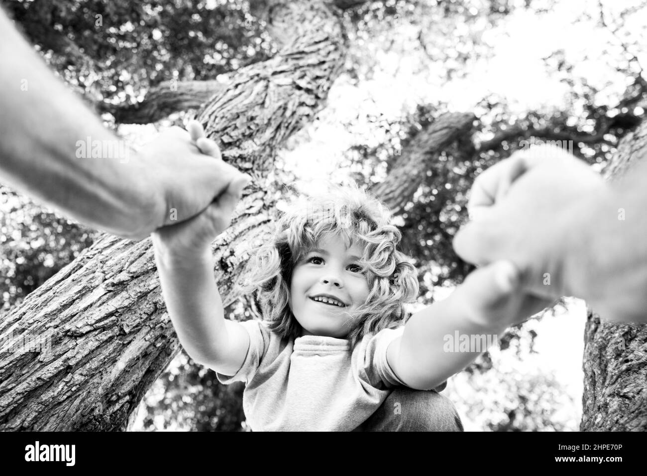 Happy boy climbing a tree during summer time. Fathers hand. Stock Photo