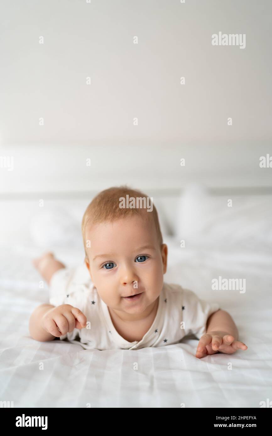 6 month old boy lies on the bed with white clothes on his stomach and laughs, looks at the camera, baby morning, happy childhood, baby products concep Stock Photo