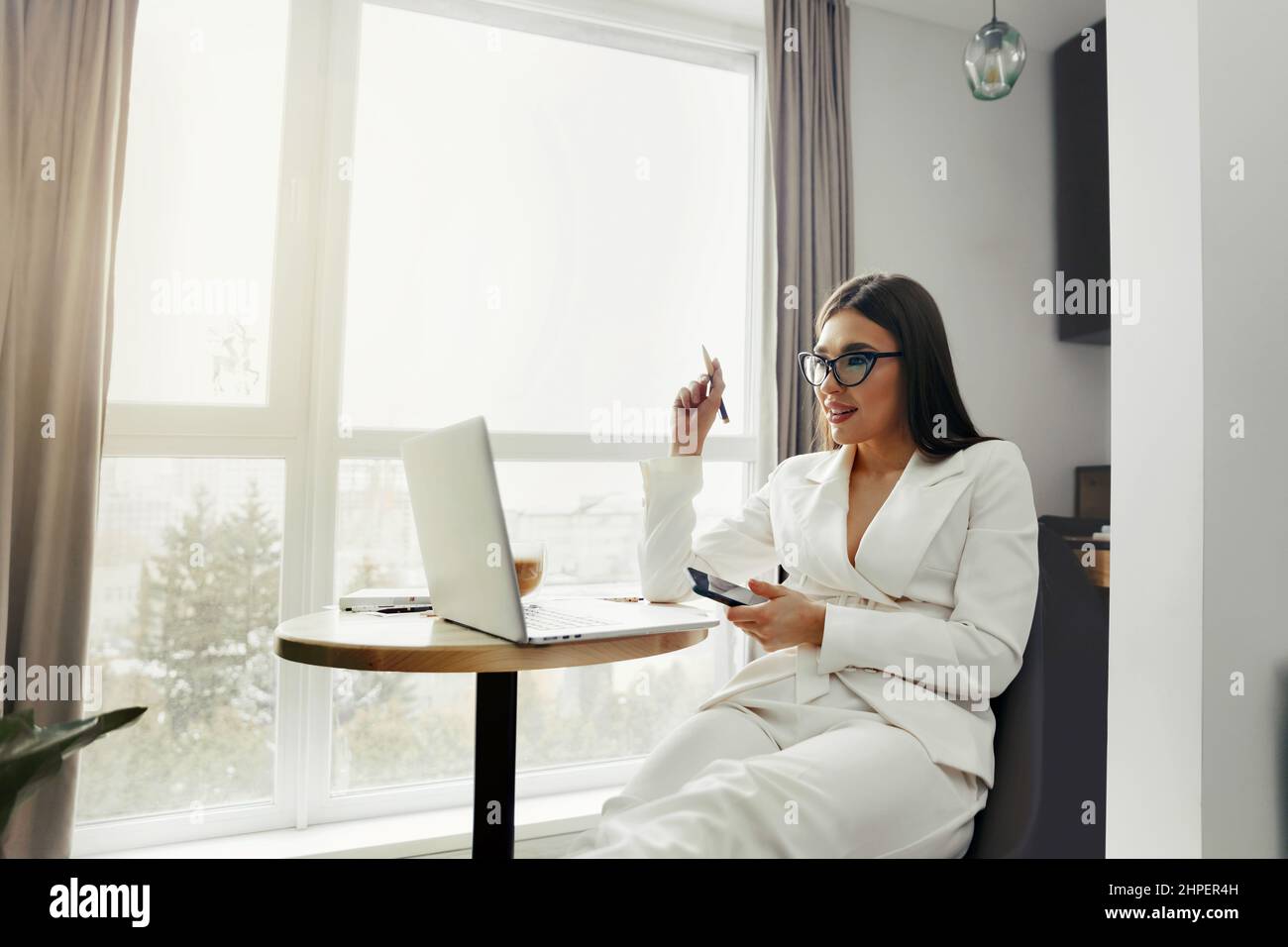 Beautiful happy woman using mobile phone while working at home with laptop. Smiling woman wearing eyeglasses messaging with smartphone. Stock Photo
