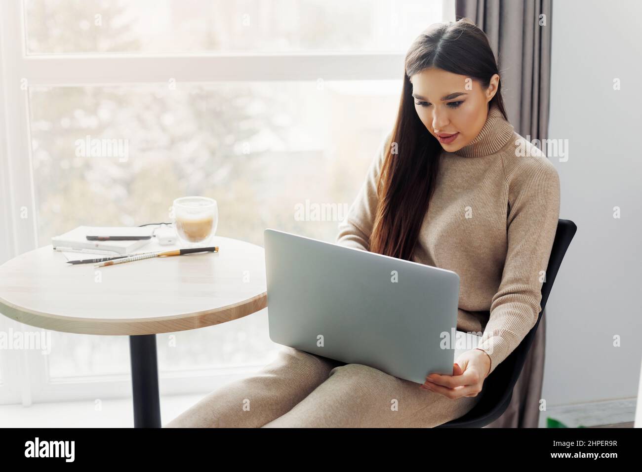 Beautiful young woman working on laptop computer while sitting at the living room, drinking coffee. Stock Photo