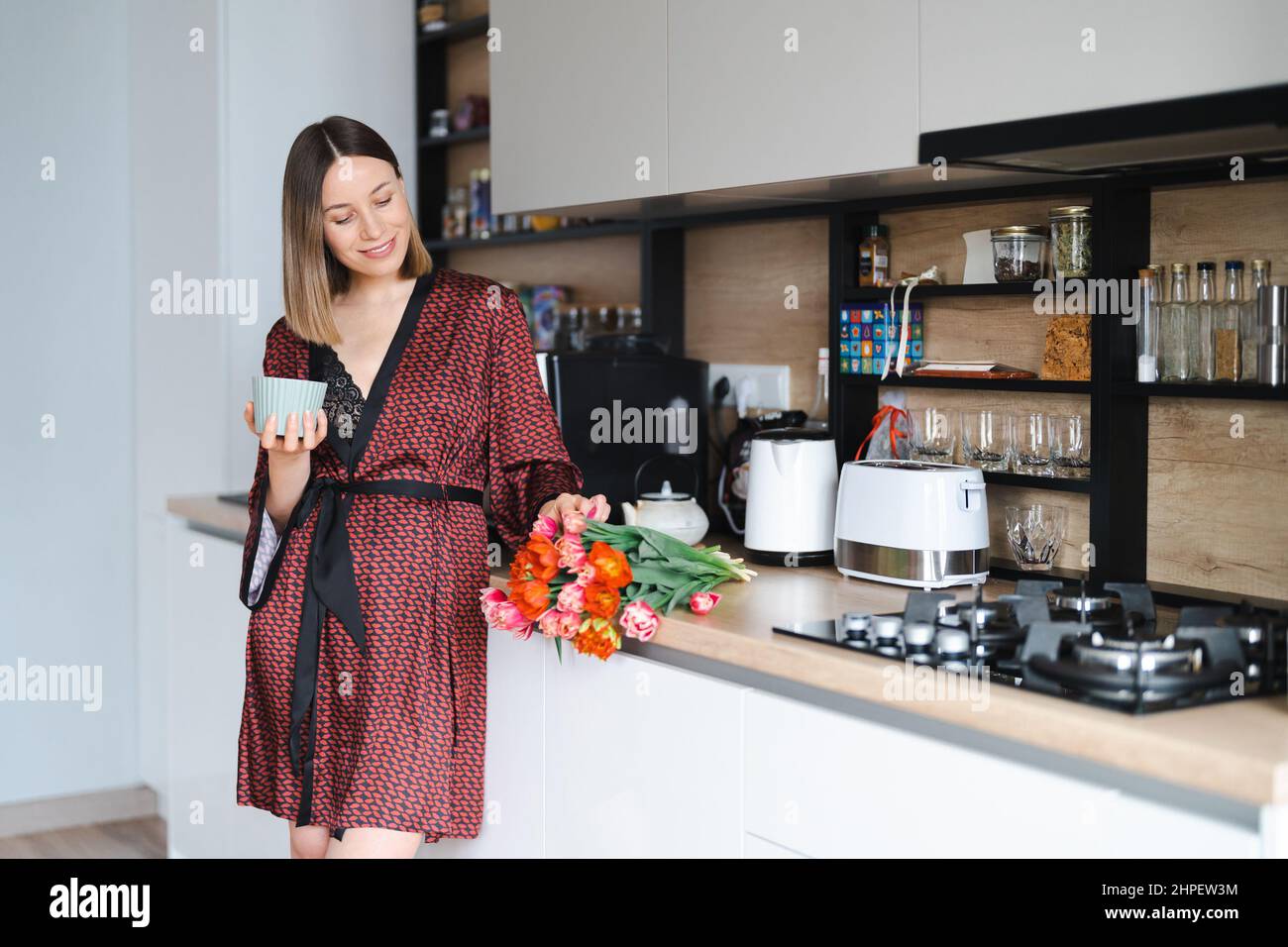 Happy Woman having a coffee at home in the kitchen wearing a silk robe  while enjoying fresh flowers Stock Photo - Alamy