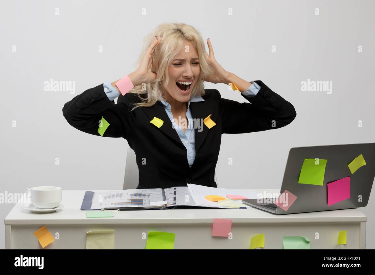 Irritated female office worker covered in sticky papers, got trouble deadline, looking at laptop and touching head Stock Photo
