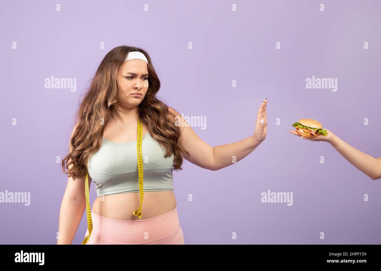 Sad plus size caucasian millennial woman in sportswear with measuring tape refuse from offered burger Stock Photo