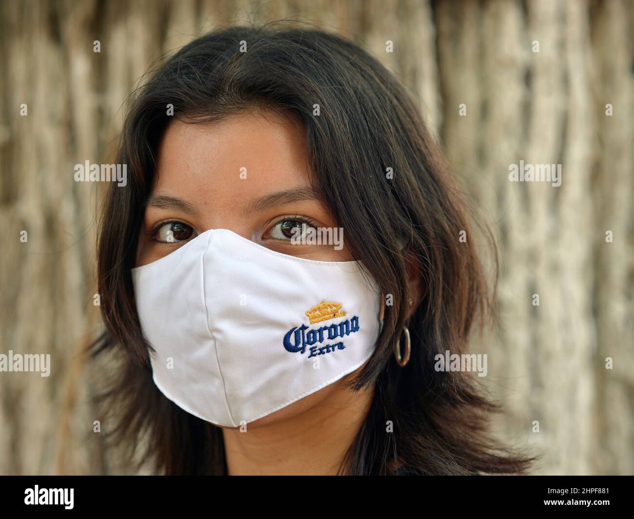 Pretty brown-eyed Mexican Latina Hispanic girl with long hair wears a white cloth face mask with corona extra beer embroidery and looks at viewer. Stock Photo