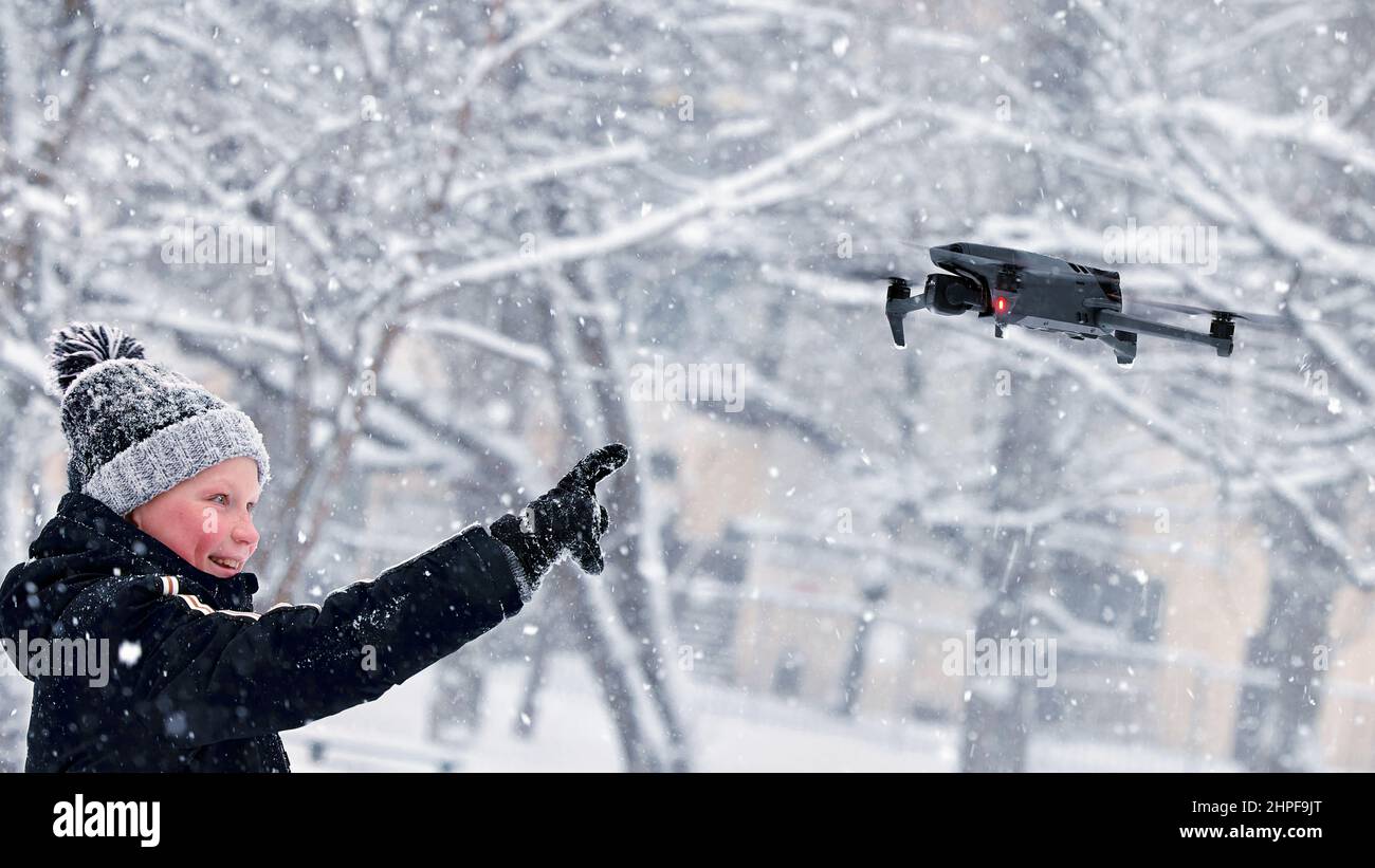 Teenager boy dressed winter jacket and modern digital drone DJI Mavic, flying in snow conditions. DJI Mavic 3 most portable drones with Hasselblad cam Stock Photo