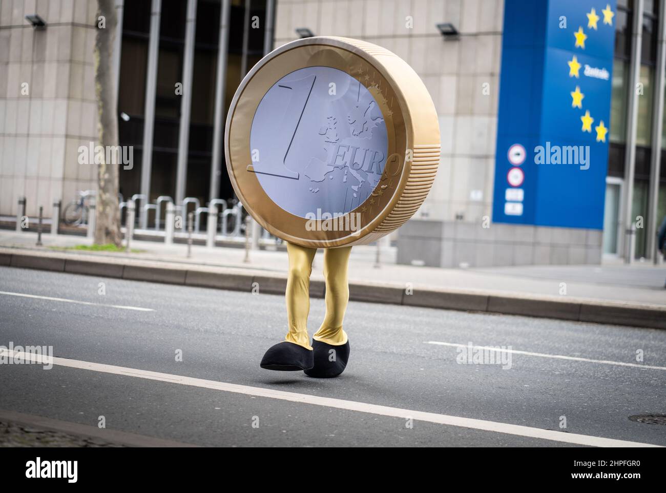 21 February 2022, Hessen, Frankfurt/Main: A performer dressed as a 'Eurolino' in the form of a one-euro coin on legs crosses a street in front of the temporary headquarters of the Deutsche Bundesbank in Frankfurt during filming. Since the actual headquarters is currently being renovated and remodeled, the Bundesbank staff and their offices are relocated to a building in the city center. The Eurolino is involved in internal filming to communicate the new locations and procedures. Photo: Frank Rumpenhorst/dpa Stock Photo