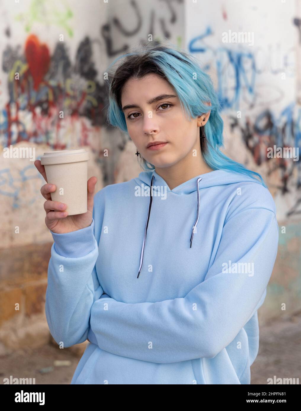 Teenage girl in light blue oversize hoodie with coffee to go and looking into the cameraBlue haired teen girl staying outdoors near graffiti wall. Clo Stock Photo