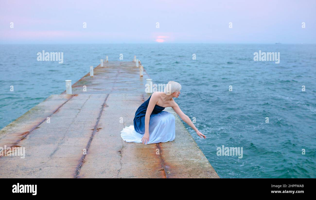 Caucasian beautiful stylish blonde woman in blue and white sitting near the sea at the sunset - protect water, save nature Stock Photo