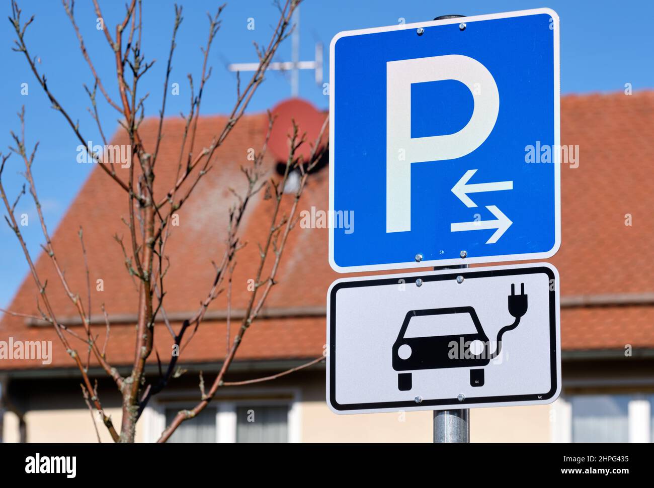 Traffic sign for a parking space for electric vehicles with charging station in front of the roof of a house. Seen in Germany in Haimendorf on a sunny Stock Photo
