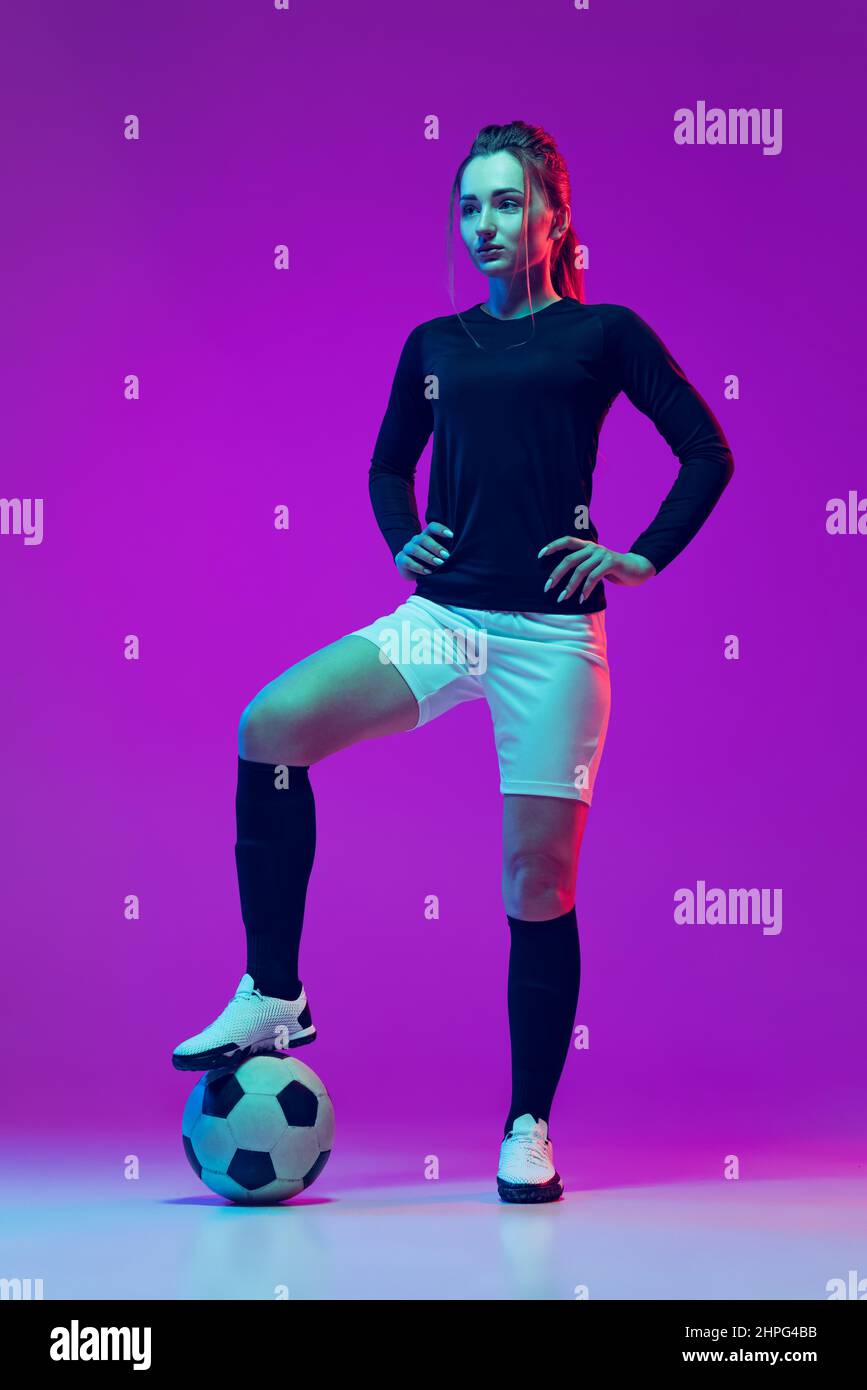 Portrait of beautiful girl, professional female soccer player posing with football ball isolated on purple studio background in neon light. Sport Stock Photo