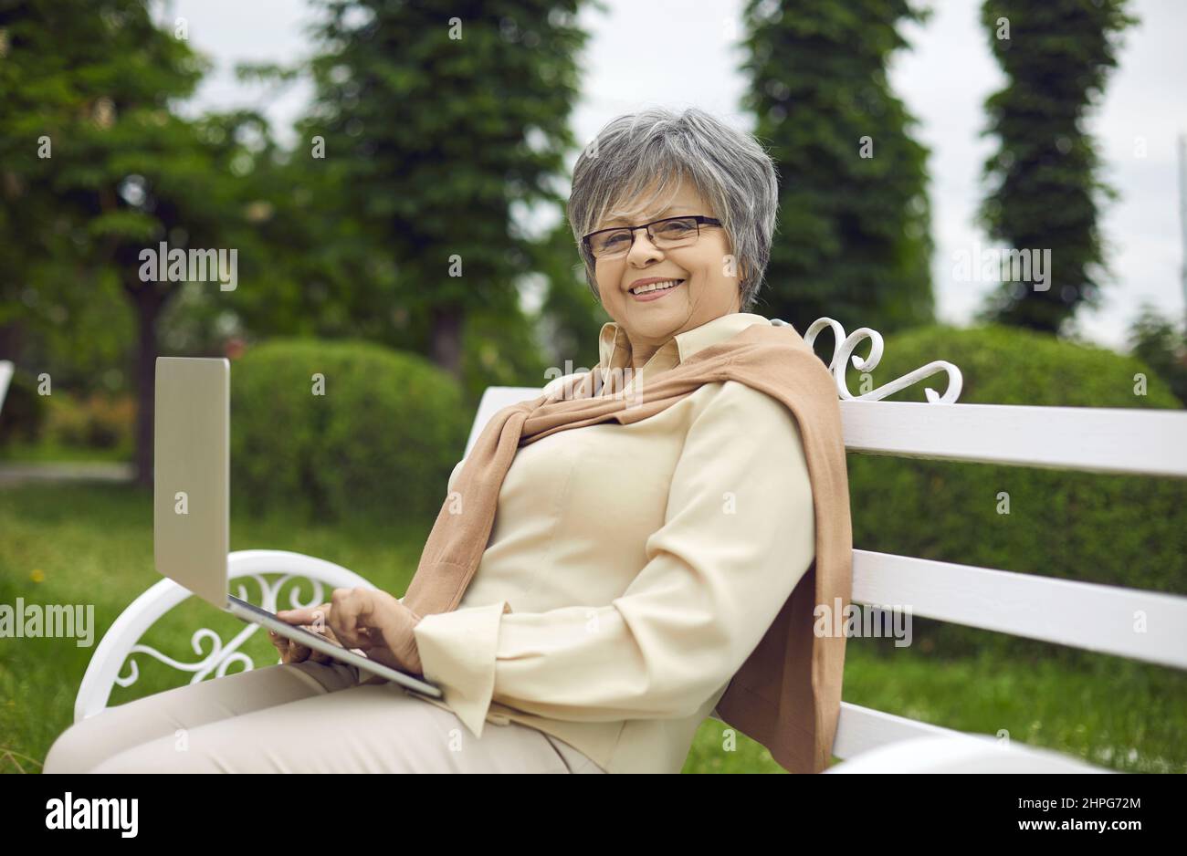 Happy elderly woman with laptop sitting on park bench looking at camera Stock Photo