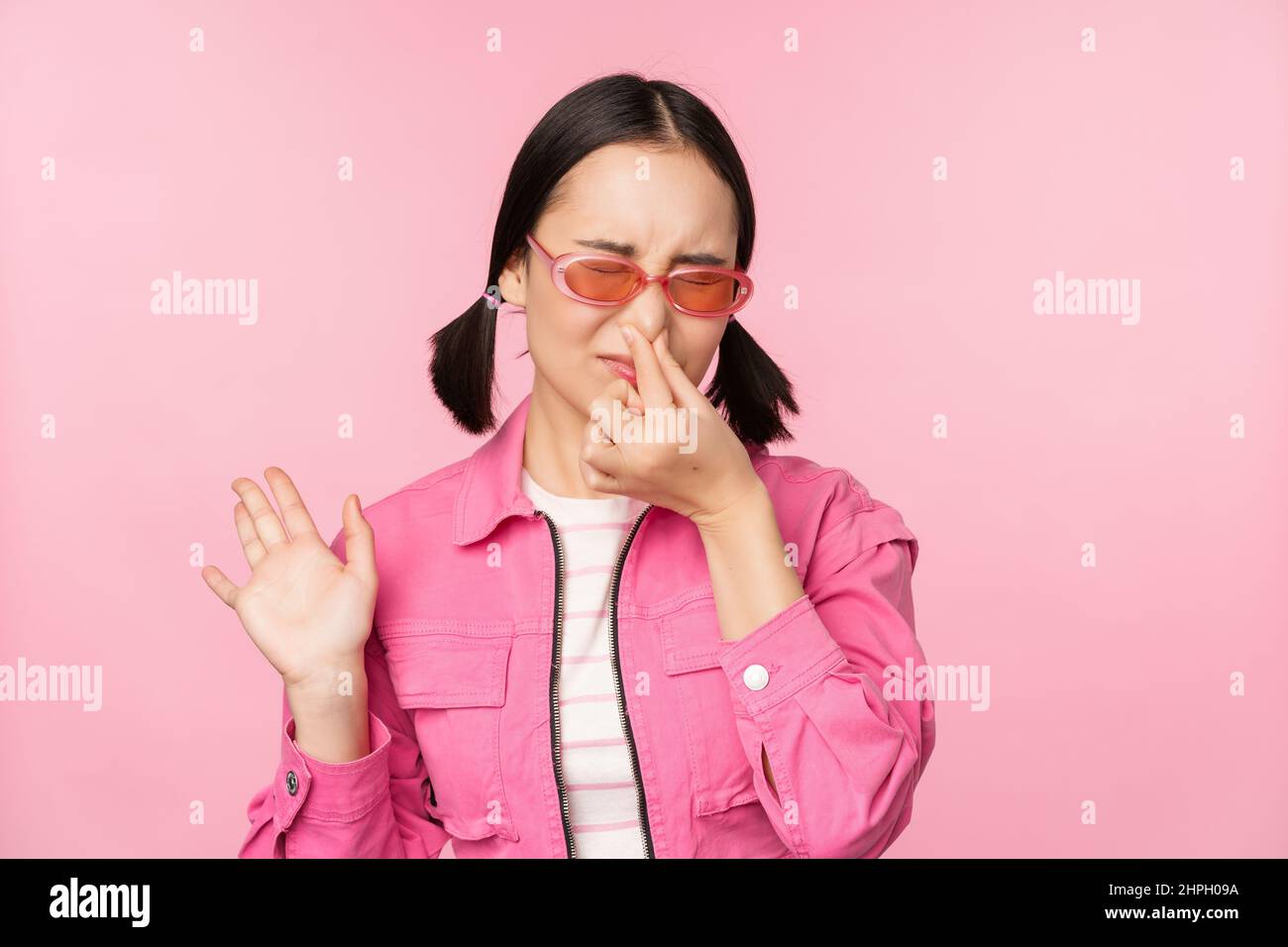 Stinky. Asian girl in sunglasses and stylish outfit, shuts her nose from disgust, dislike bad smell, standing over pink background Stock Photo