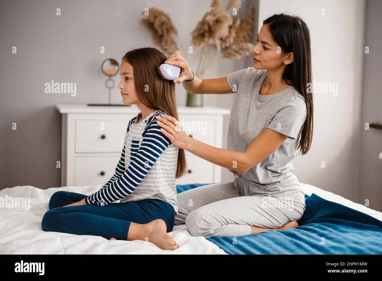 Beautiful young mother combing her cute little daughters hair while sitting on bed and smiling Stock Photo