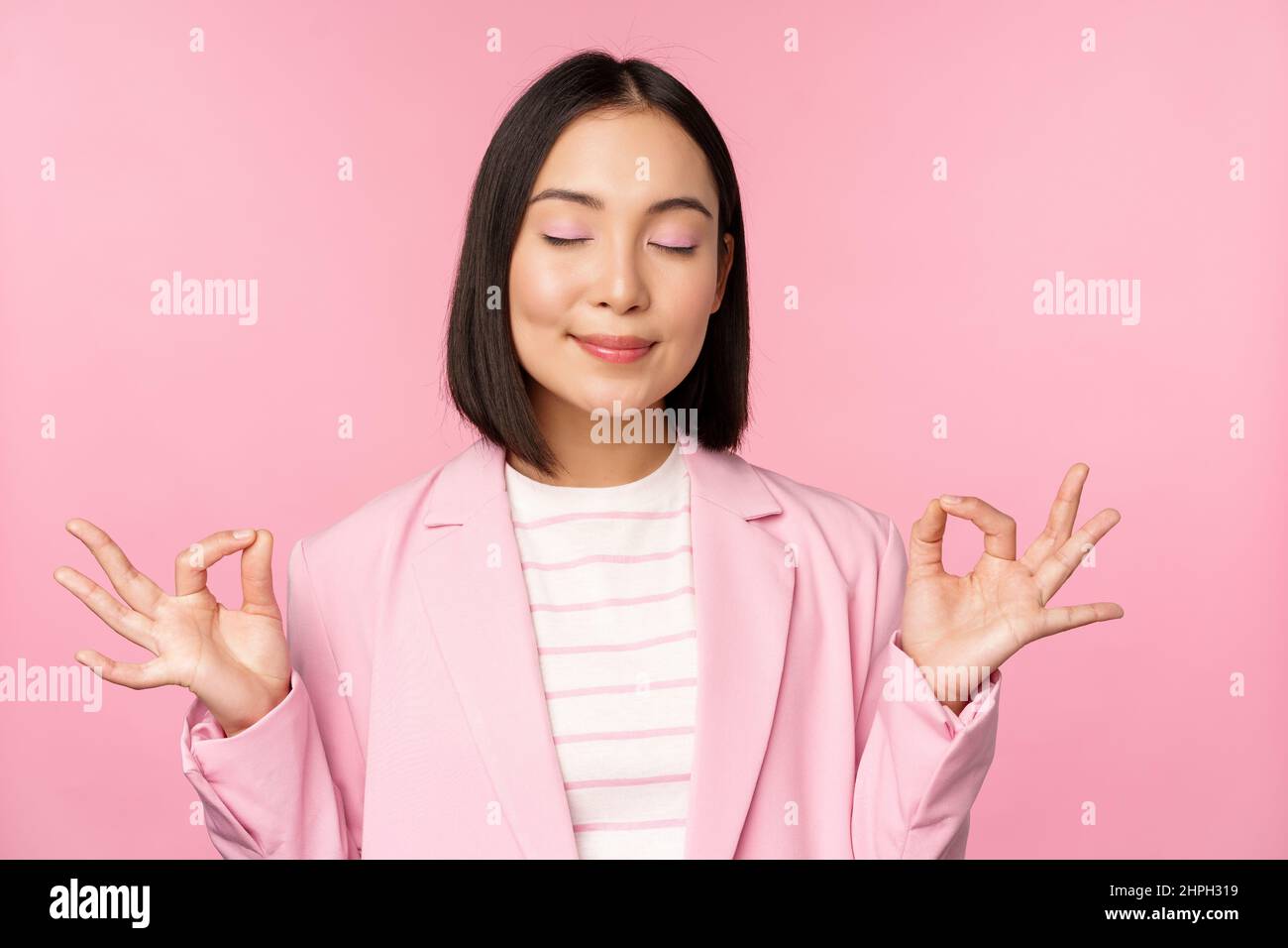 Keep calm. Smiling young asian woman meditating, practice yoga, mindfulness and relaxation at work, breath, inhale air with pleased face, standing Stock Photo