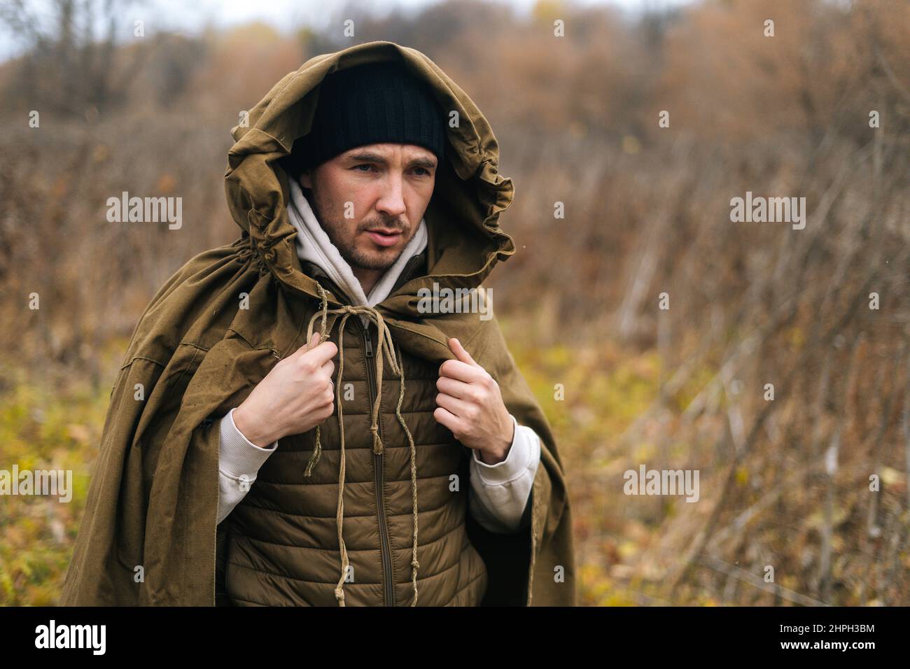Portrait of thoughtful tourist man wearing green raincoat tent standing in thicket of bushes in cold overcast day and looking away. Stock Photo