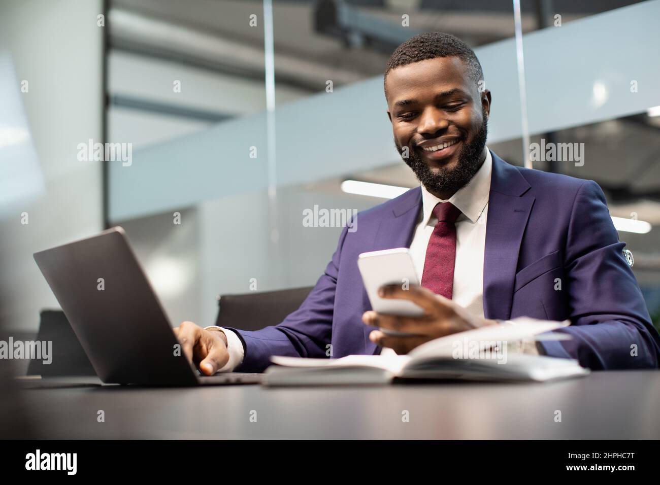 Cheerful black manager using smartphone while working on laptop Stock Photo